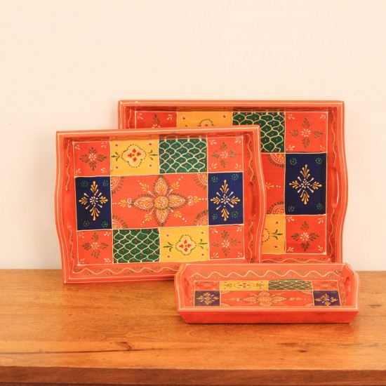 Set of three nesting wooden serving trays in orange, hand painted in a colourful patchwork pattern of floral and geometric motifs in green, blue, and yellow, with cut-out side handles.