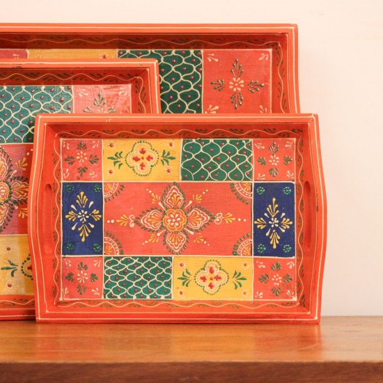 Set of three nesting wooden serving trays in orange, hand painted in a colourful patchwork pattern of floral and geometric motifs in green, blue, and yellow, with cut-out side handles.