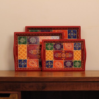 Set of three nesting wooden serving trays in red, hand painted in a colourful patchwork pattern of floral and geometric motifs in green, blue, and yellow, with cut-out side handles.