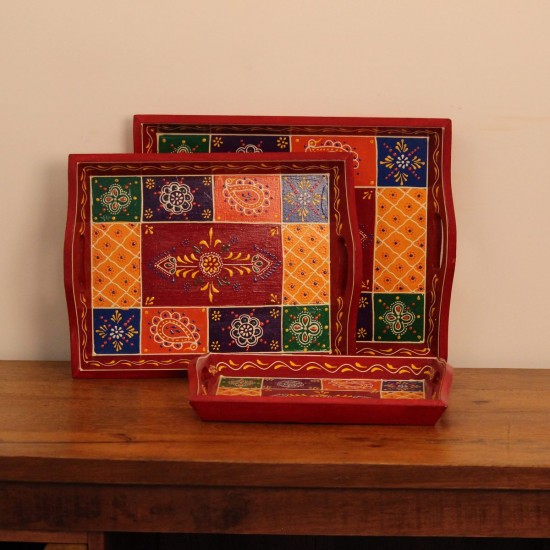 Hand Painted Red Patchwork Tray – Set of 3 Set of three nesting wooden serving trays in red, hand painted in a colourful patchwork pattern of floral and geometric motifs in green, blue, and yellow, with cut-out side handles.