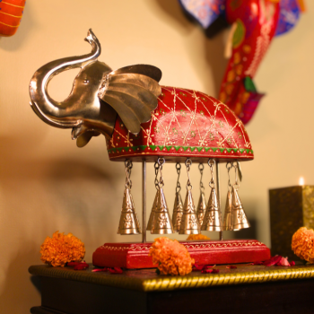 Hand-painted red Bell Elephant statuette with upraised trunk and ten metal bells on a wooden stand, styled with marigold garlands and a lit diya — traditional Rajasthani pooja décor (wood & metal).