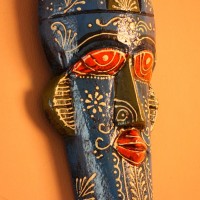 Hand-carved wooden tribal face mask hand painted in blue with a mustard-yellow crown, red eyes and lips, and fine white floral detailing across the face.