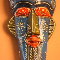 Hand-carved wooden tribal face mask hand painted in blue with a mustard-yellow crown, red eyes and lips, and fine white floral detailing across the face.