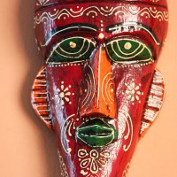 Hand Painted Tribal Face Mask – Maroon Floral Hand-carved wooden tribal face mask hand painted in deep maroon with green eyes and lips, a blue feathered crown, and fine white and orange floral motifs across the face.