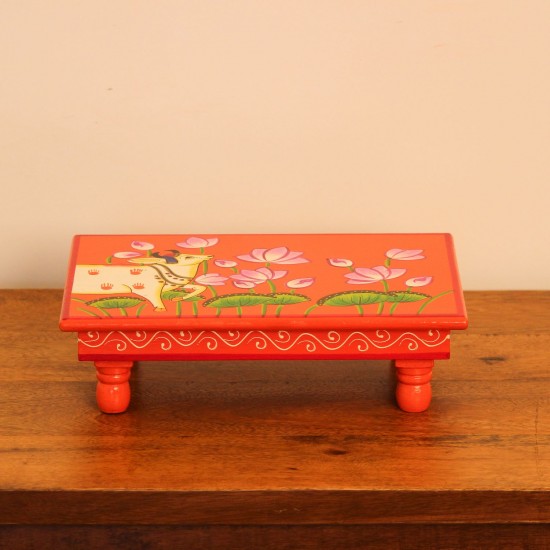 Hand Painted Cow & Lotus Chowki – Large Large wooden chowki hand painted in Pichwai style, featuring a cow standing amid blooming pink lotus flowers on a vivid red base, edged with a plain wooden border.