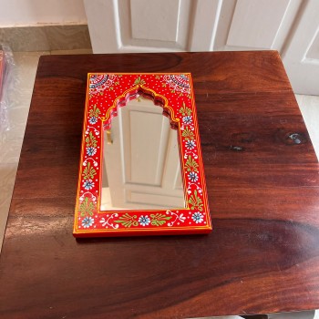 Hand-carved wooden mirror frame in a jharokha arch shape, hand painted in red with a fine floral border in blue, green, and white, framing an arched mirror at the centre.