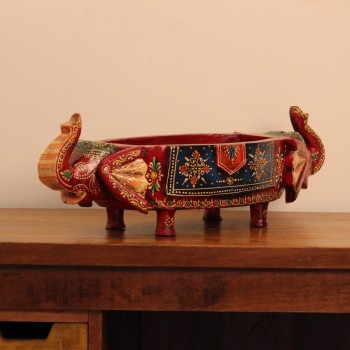Wooden fruit bowl flanked by two carved elephant heads with brass tusk accents, hand painted in red with a blue floral panel across the bowl's front.
