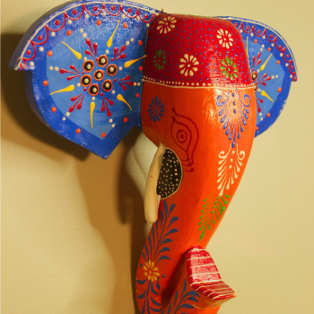 Hand-painted wooden elephant face wall decor in blue and orange placed above a traditional Indian decor setup