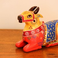 Multicolour Nandi Figurine Styled on Table Hand-painted multicolour wooden Nandi figurine with an orange-red body placed on a wooden table as a pooja or console accent.