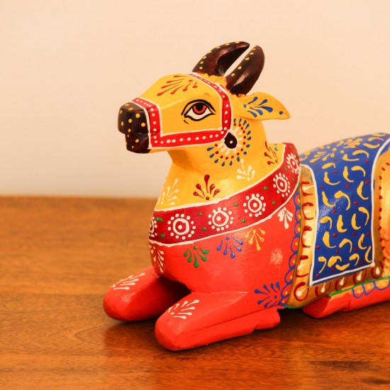 Multicolour Nandi Figurine Styled on Table Hand-painted multicolour wooden Nandi figurine with an orange-red body placed on a wooden table as a pooja or console accent.