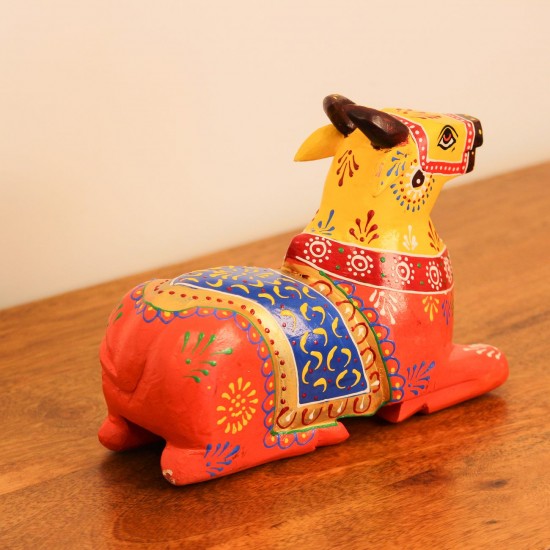 Multicolour Nandi Figurine Styled on Table Hand-painted multicolour wooden Nandi figurine with an orange-red body placed on a wooden table as a pooja or console accent.