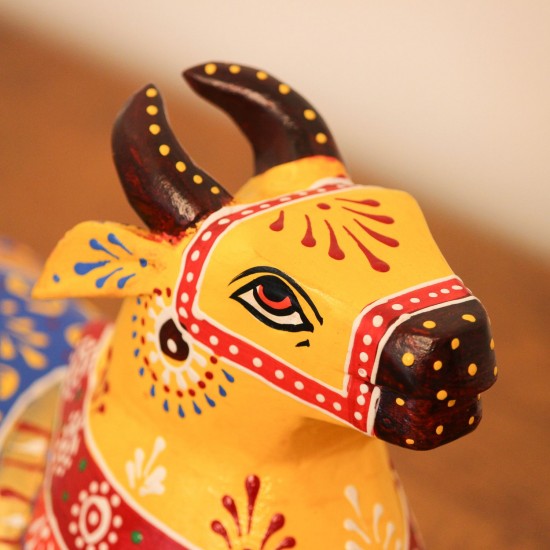 Multicolour Nandi Figurine Styled on Table Hand-painted multicolour wooden Nandi figurine with an orange-red body placed on a wooden table as a pooja or console accent.