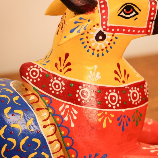 Multicolour Nandi Figurine Styled on Table Hand-painted multicolour wooden Nandi figurine with an orange-red body placed on a wooden table as a pooja or console accent.
