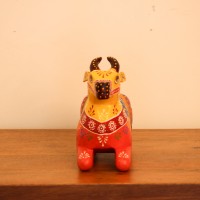 Multicolour Nandi Figurine Styled on Table Hand-painted multicolour wooden Nandi figurine with an orange-red body placed on a wooden table as a pooja or console accent.
