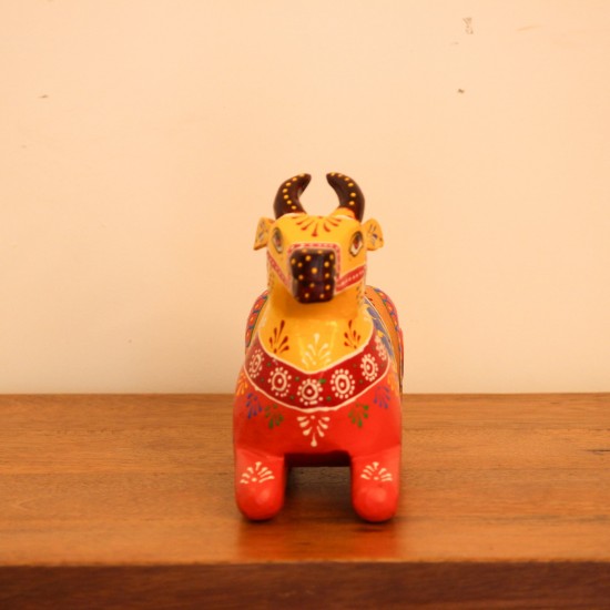 Multicolour Nandi Figurine Styled on Table Hand-painted multicolour wooden Nandi figurine with an orange-red body placed on a wooden table as a pooja or console accent.