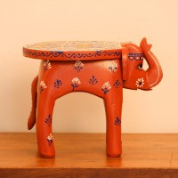 Hand Painted Burnt Orange Elephant Stool Styled on Table Hand-painted burnt orange-brown wooden elephant stool placed on a wooden table as a side stool or display stand.